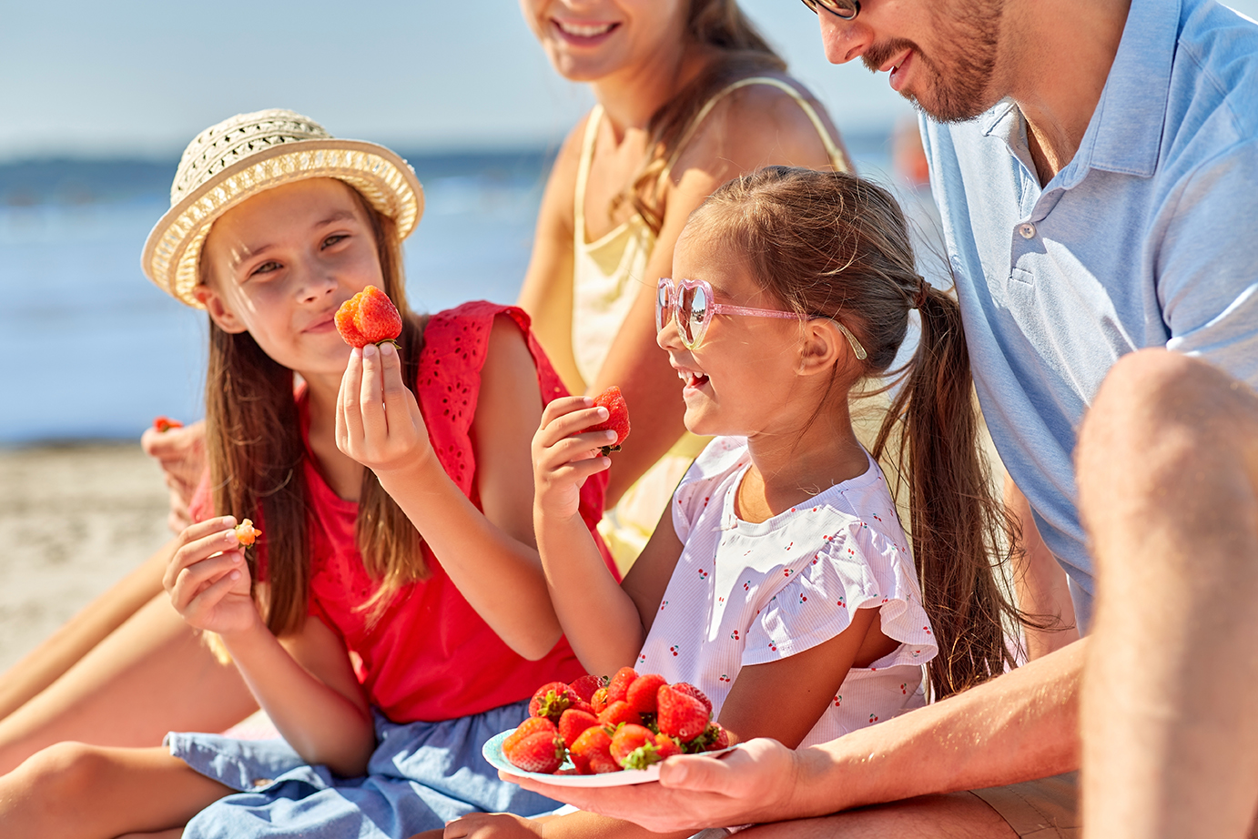 family, happy mother, father and two daughters having picnic on summer beach and eating strawberries