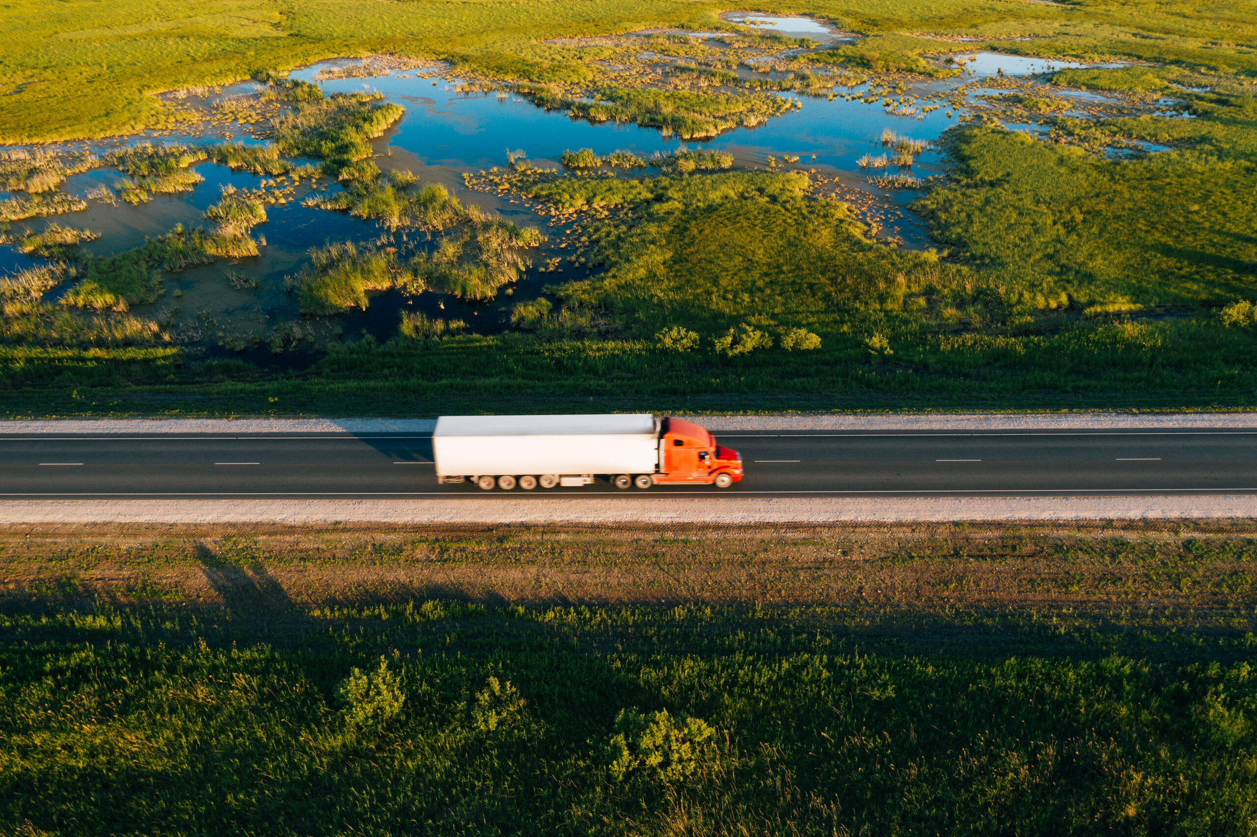 Semi truck with cargo trailer drive on road in green grass field with water at sunset, aerial drone view. 