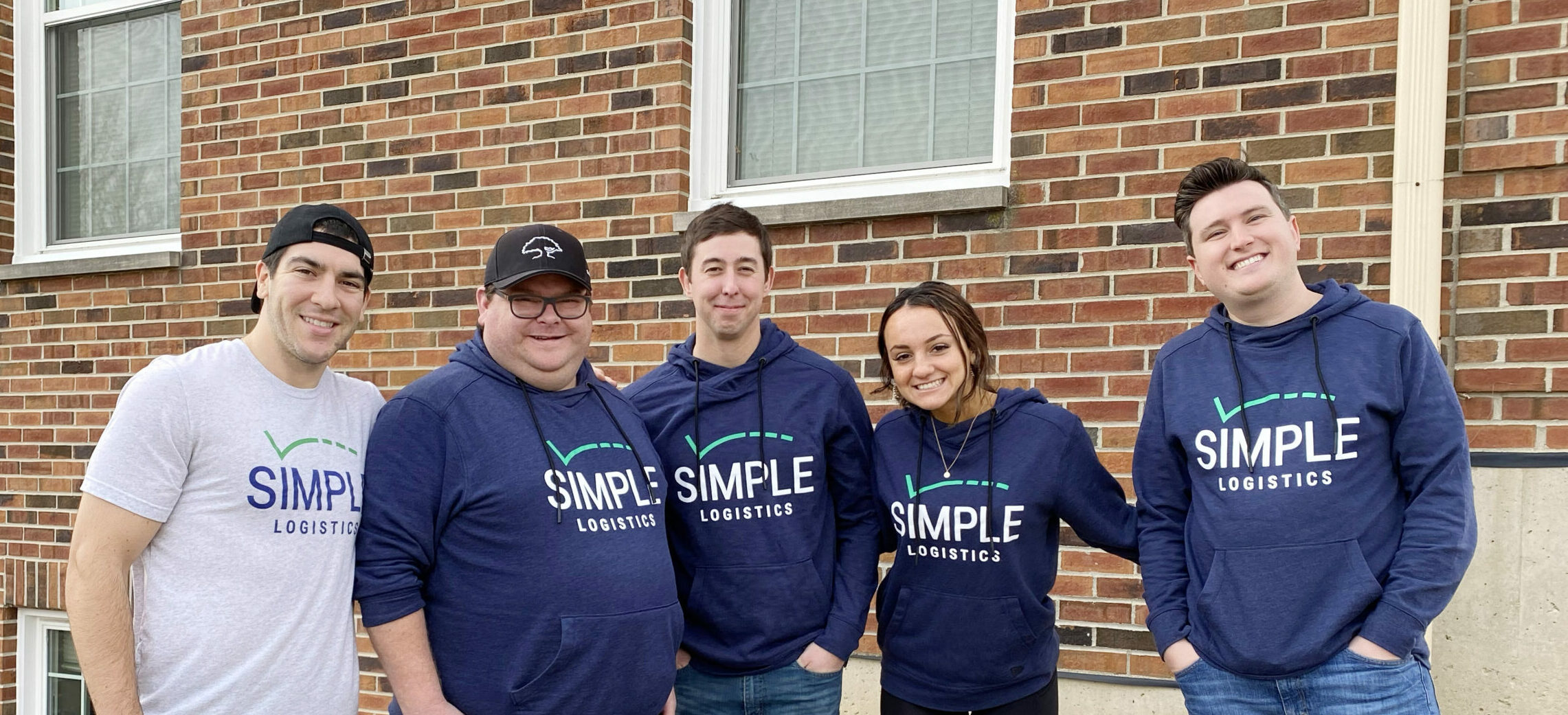 5 Members of the Simple Logistics Team standing together in front of a brick building, smiling at the camera