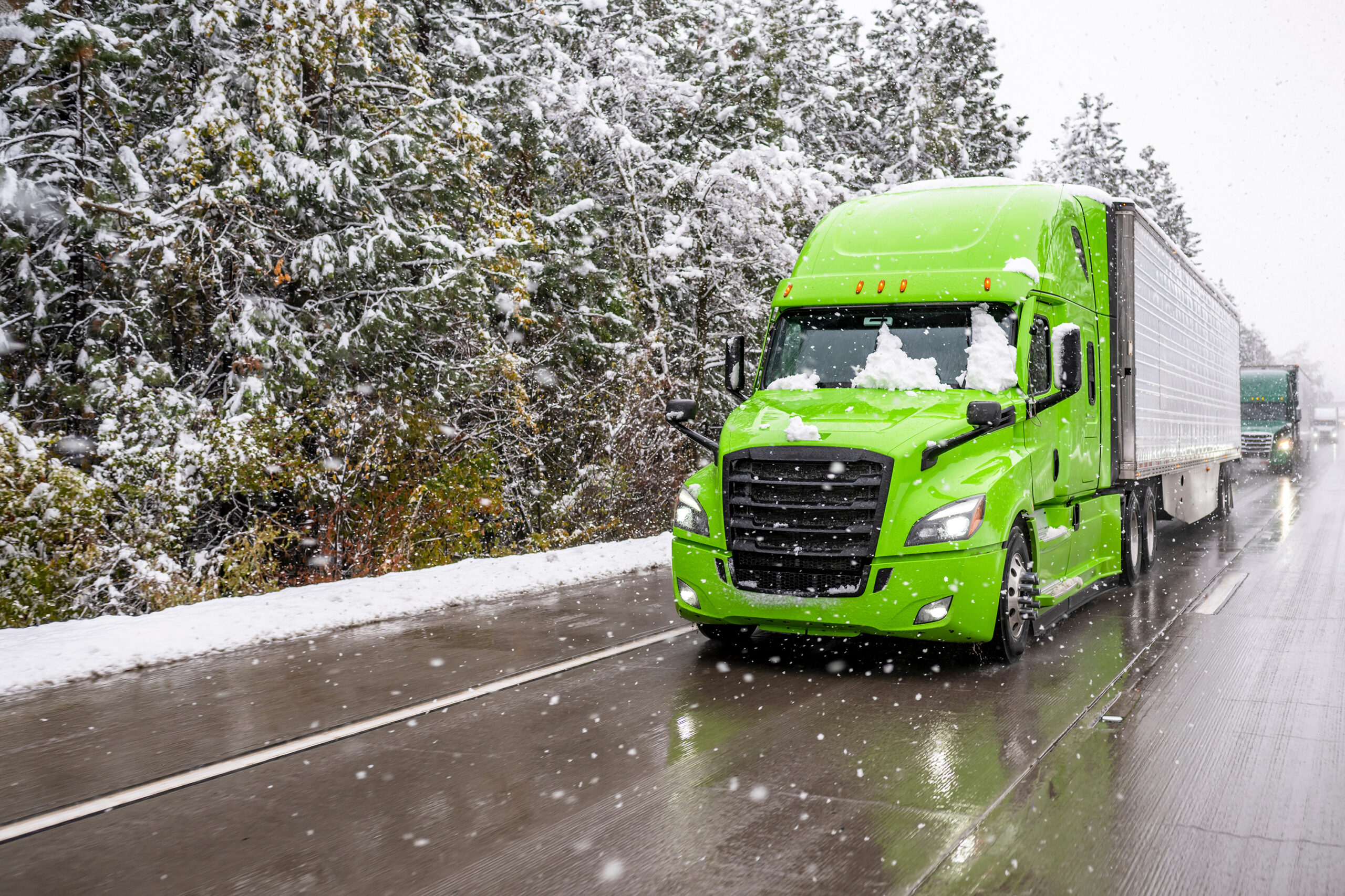Green semi-truck transporting cargo in refrigerator semi-trailer driving in front of convoy moving slowly on winter highway during a snowstorm.